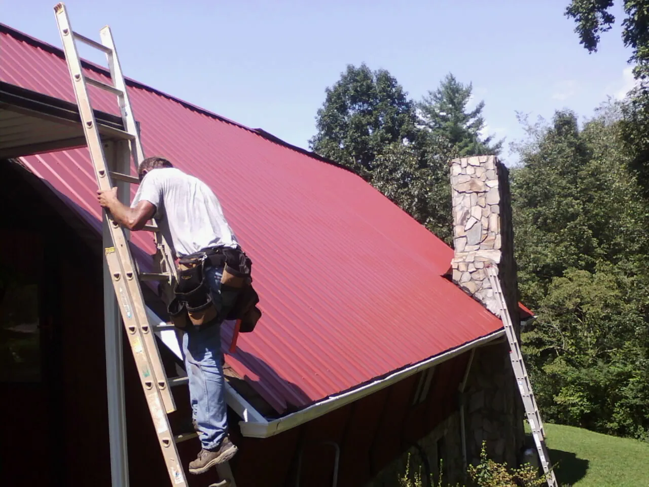 Roofer on a ladder beside a red metal roof and stone chimney — Soffit & Fascia Repair by Roofing Brothers in Dagsboro